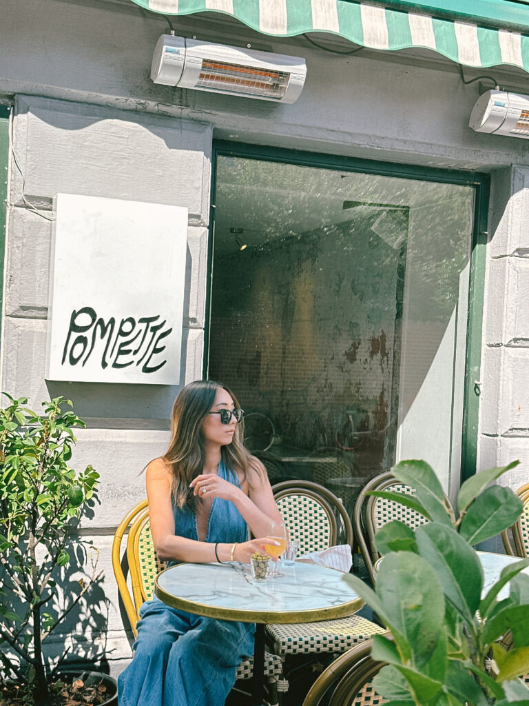 Woman with sunglasses seated at an outdoor cafe table with a drink, leafy plants in foreground and a pale building behind her.