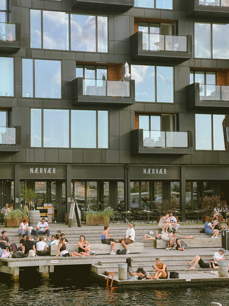 People lounging on concrete steps by a canal in front of a dark, modern building with balconies and large windows.