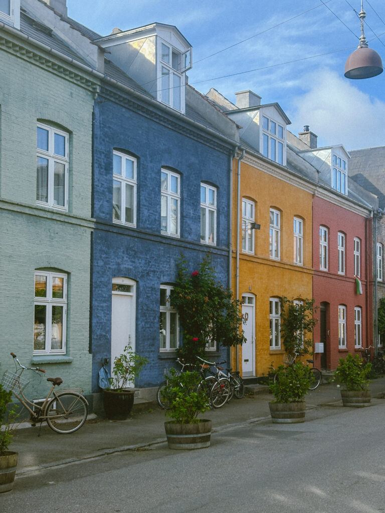 Colorful row houses in pastel green, blue, yellow, and pink lining a street with bicycles and potted plants on the sidewalk under a blue sky.