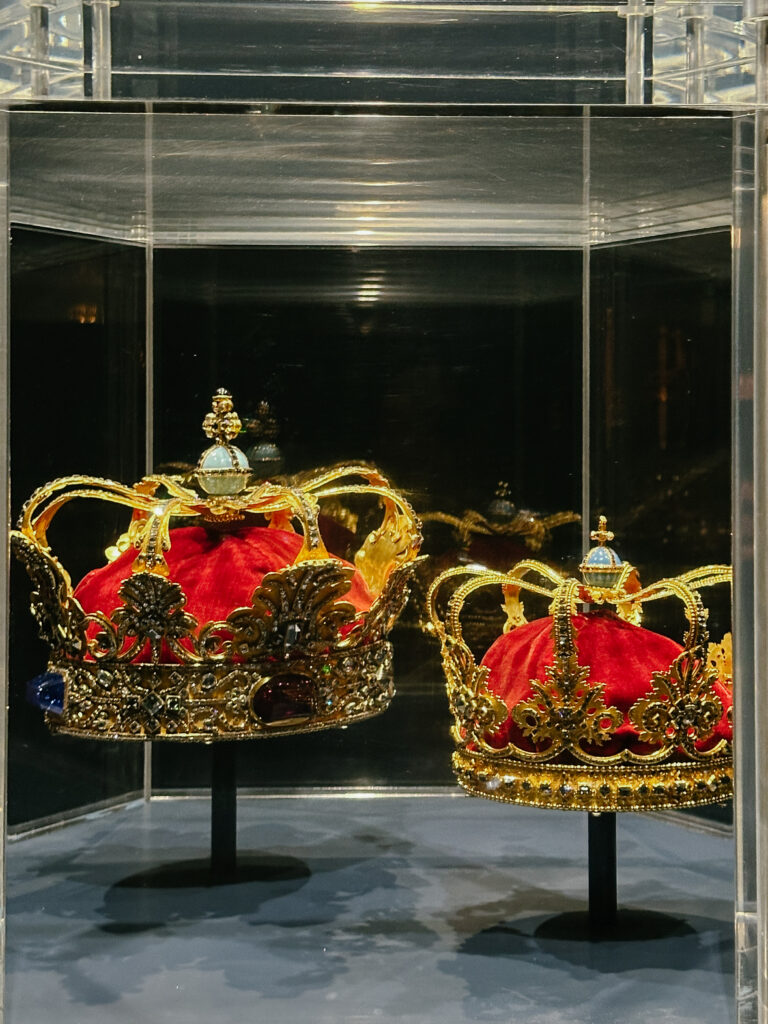 Two ornate gold crowns encrusted with gems resting on red velvet cushions inside a glass display case against a dark backdrop.