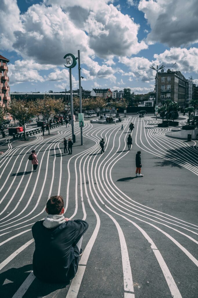 Wide city plaza with white curved lines on the pavement, pedestrians walking, and a blue, cloud-filled sky above.