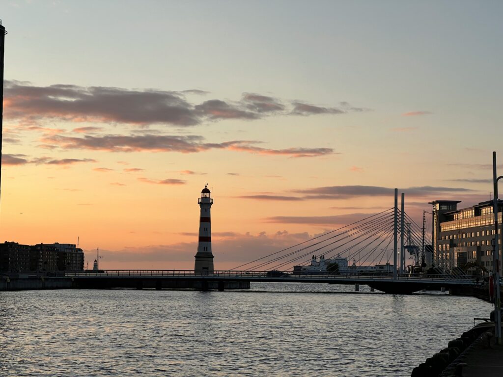 Sunset over a harbor with a striped lighthouse in the foreground and a cable-stayed bridge to the right.