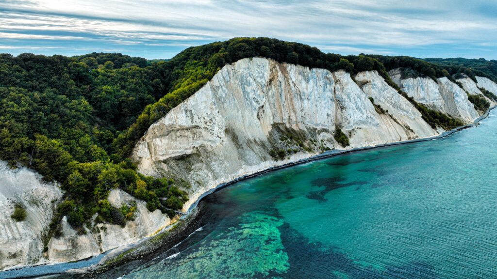White chalk cliffs meet a forested coastline and turquoise sea below, under a blue sky.