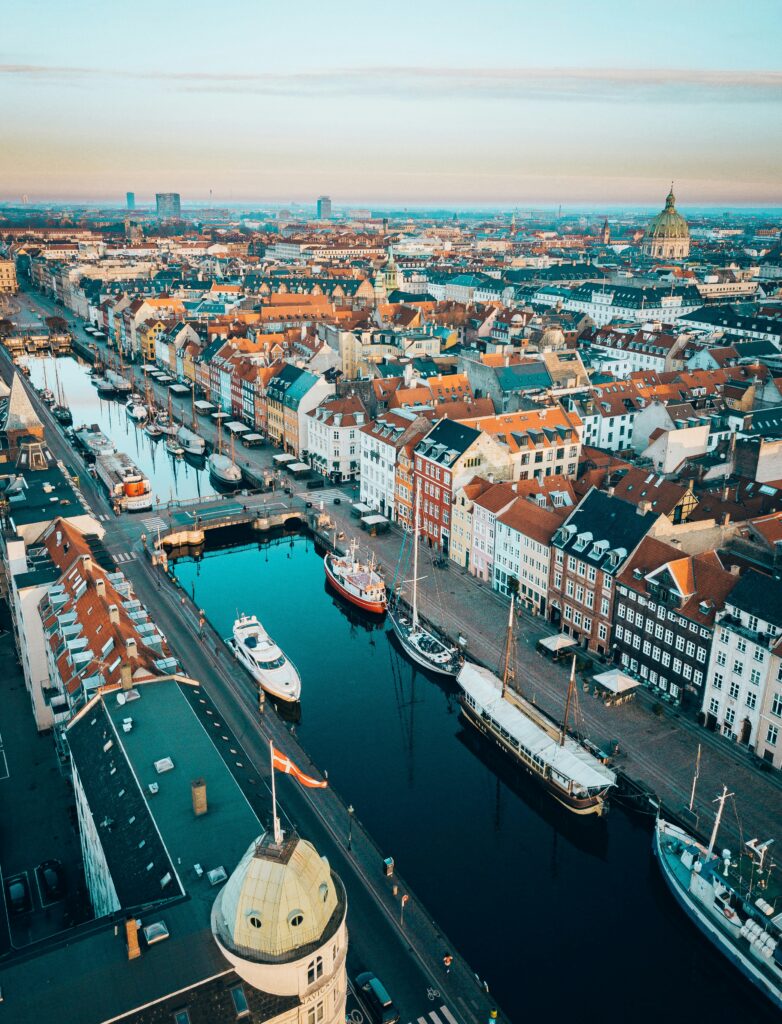 Aerial view of a European harbor city: canal with moored boats, colorful row houses, and a large domed church in the distance.
