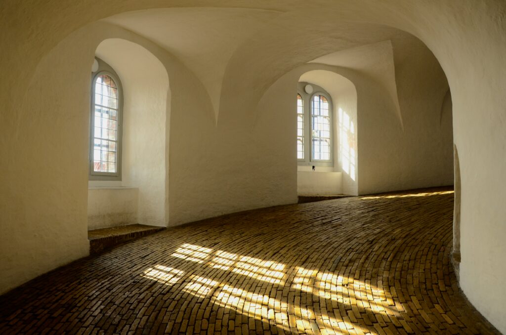 Sunlit interior with curved white walls, arched windows, and a brick floor forming striped light patterns on the ground.